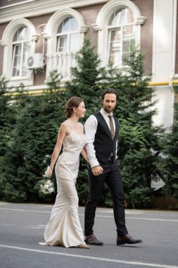 full length of groom walking with happy bride in white dress holding bouquet of flowers 