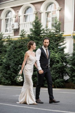 full length of groom walking with happy bride in white dress holding bouquet of flowers  clipart