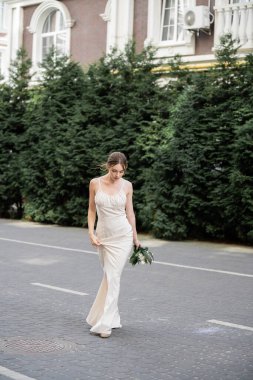 full length of young bride in white dress holding wedding bouquet of flowers and walking on street 