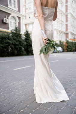 partial view of young woman in white dress holding wedding bouquet behind back  clipart