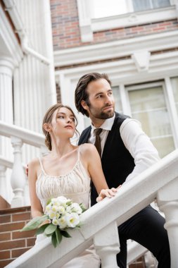 Gelinlik giymiş genç bir kadın ve takım elbiseli bir damat evin yakınına bakıyor. 
