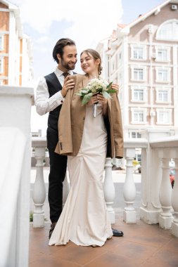 full length of happy groom in vest hugging cheerful bride in beige blazer and wedding dress with bouquet 