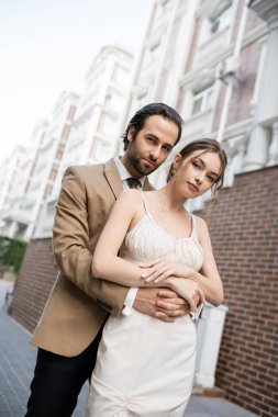 groom in beige suit hugging gorgeous bride in white wedding dress 