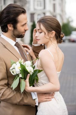 pretty bride in wedding dress holding blooming flowers and standing with groom 