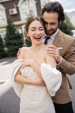 portrait of cheerful and bearded man hugging happy bride laughing with closed eyes