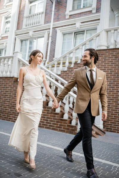 full length of happy newlyweds smiling and holding hands while walking on street
