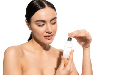 brunette woman with bare shoulders looking at bottle with moisturizing serum isolated on white 