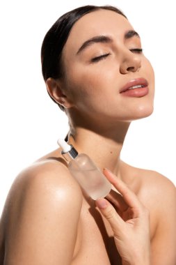 brunette woman with bare shoulders and closed eyes holding bottle with moisturizing serum isolated on white 