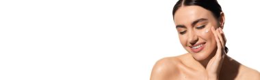 satisfied young woman applying moisturizing face cream on cheek isolated on white, banner 