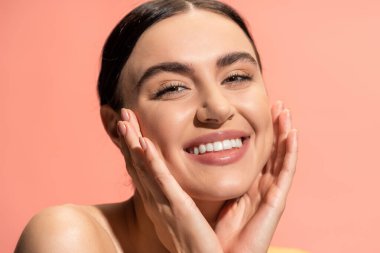 pleased young woman with bare shoulders touching soft cheeks isolated on pink 