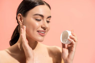 portrait of cheerful woman with bare shoulders holding jar with nourishing cream isolated on pink 
