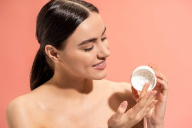 portrait of pleased woman with bare shoulders holding jar with nourishing cream isolated on pink 