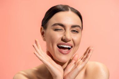 excited young woman winking and gesturing isolated on pink 