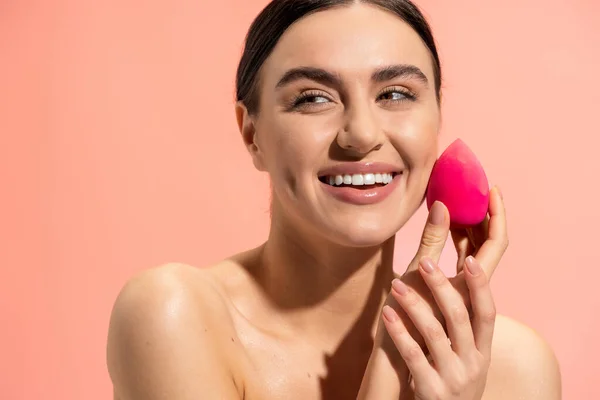 joyful woman applying face foundation with beauty sponge isolated on pink