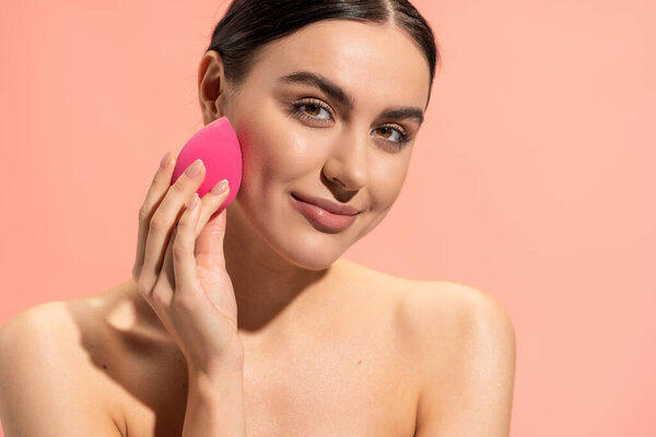 happy woman with bare shoulders applying face foundation with beauty sponge isolated on pink 