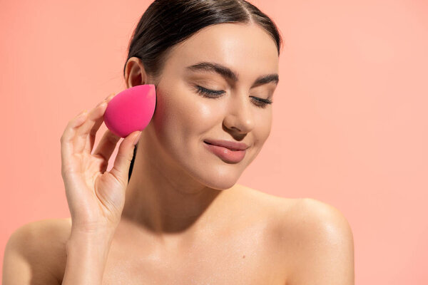 pleased woman applying face foundation with makeup sponge isolated on pink 