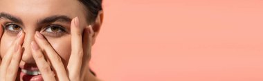 close up view of happy young woman covering face while looking at camera isolated on pink, banner 