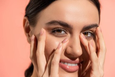 close up view of cheerful young woman touching face while looking at camera isolated on pink  clipart