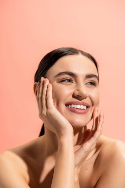 smiling woman with bare shoulders looking away while touching cheek isolated on pink 