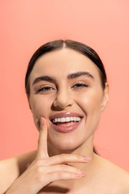charming woman smiling while touching face with finger isolated on pink 