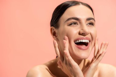 excited young woman with bare shoulders touching cheeks isolated on pink 