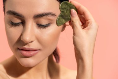 young brunette woman with soft skin doing face massage with jade scraper isolated on pink 