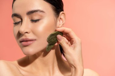 brunette woman with bare shoulders and soft skin doing jade massage with face scraper isolated on pink 