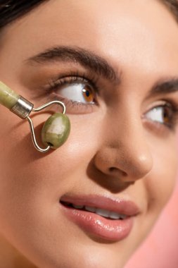 close up view of smiling woman doing face massage with jade roller isolated on pink 