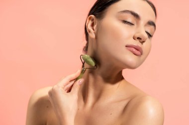 brunette young woman doing neck massage with jade roller isolated on pink  clipart