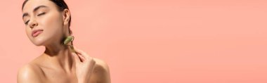brunette young woman doing neck massage with jade roller isolated on pink, banner 