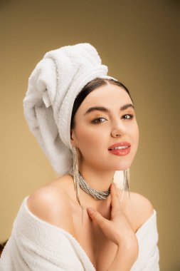 cheerful woman with makeup and towel on head looking at camera on beige background 
