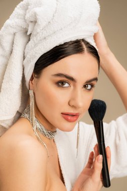 brunette woman with white towel on head holding makeup brush on grey background 