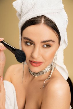 brunette woman with towel on head applying face powder with cosmetic brush on beige background 