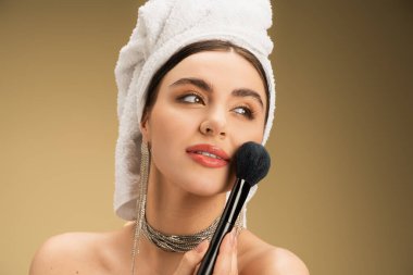 young woman with towel on head applying face powder with cosmetic brush on beige background  clipart