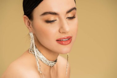 brunette woman in necklace and earrings looking away while posing isolated on beige 