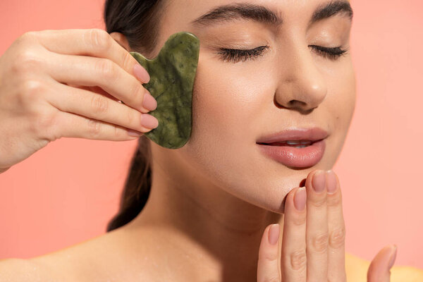 brunette woman with closed eyes doing massage with face scraper isolated on pink 