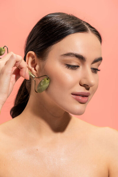 brunette young woman doing massage with jade roller isolated on pink 