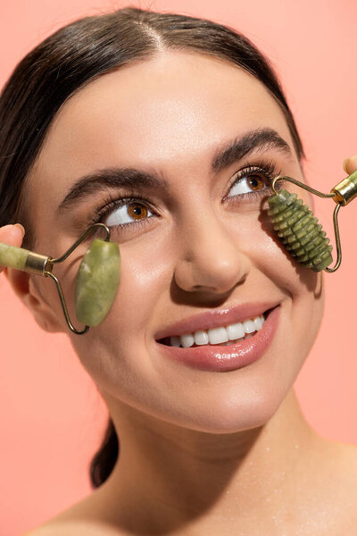 close up view of cheerful young woman doing face massage with jade rollers isolated on pink 