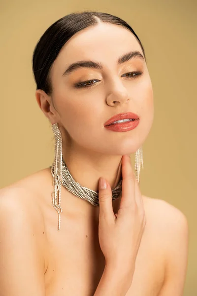 sensual woman in necklace and earrings touching neck isolated on beige