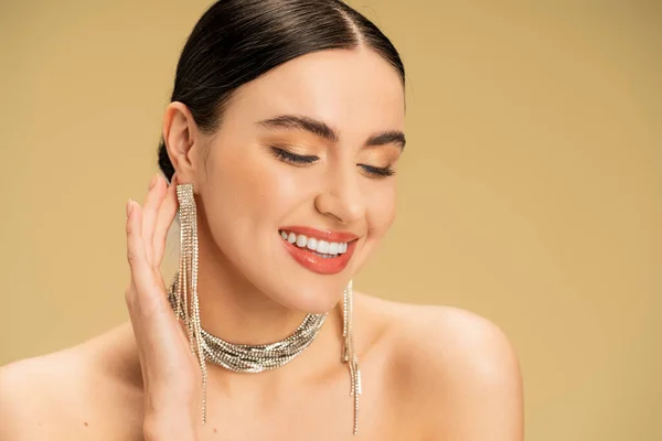brunette young woman in necklace and earrings smiling isolated on beige