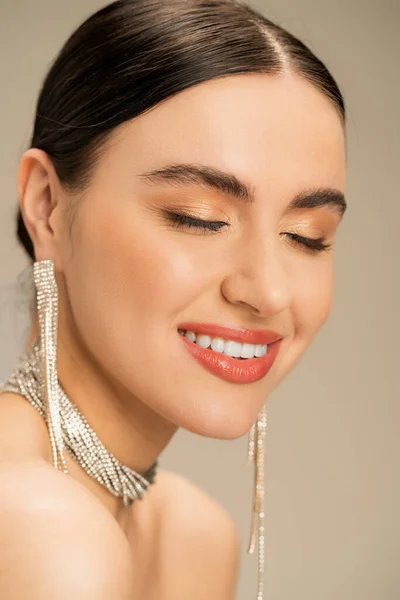 joyful young woman in necklace and earrings smiling with closed eyes isolated on beige