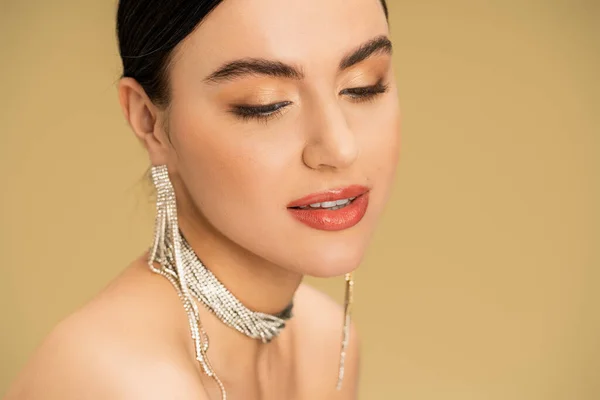 brunette woman in necklace and earrings looking away while posing isolated on beige