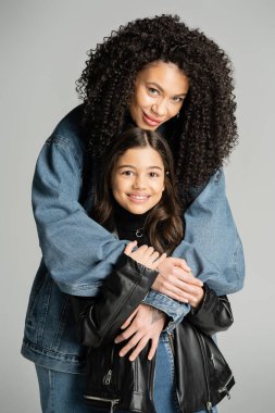 Smiling woman in denim jacket hugging daughter isolated on grey 