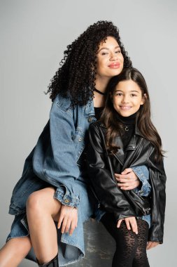 Positive woman looking at camera and hugging daughter isolated on grey 