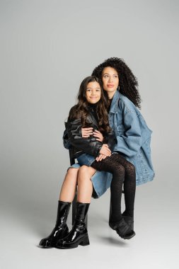 Stylish woman holding smiling daughter while sitting on grey background 