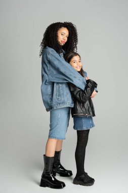 Full length of stylish brunette woman hugging happy preteen daughter on grey background 
