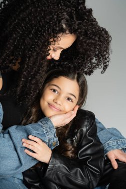 Portrait of curly mother hugging cheerful kid isolated on grey 
