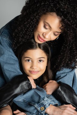 Stylish young woman hugging cheerful preteen daughter isolated on grey 
