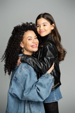 Smiling curly mother in denim jacket hugging stylish child isolated on grey 