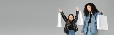 Cheerful preteen girl holding shopping bags near mom isolated on grey, banner 